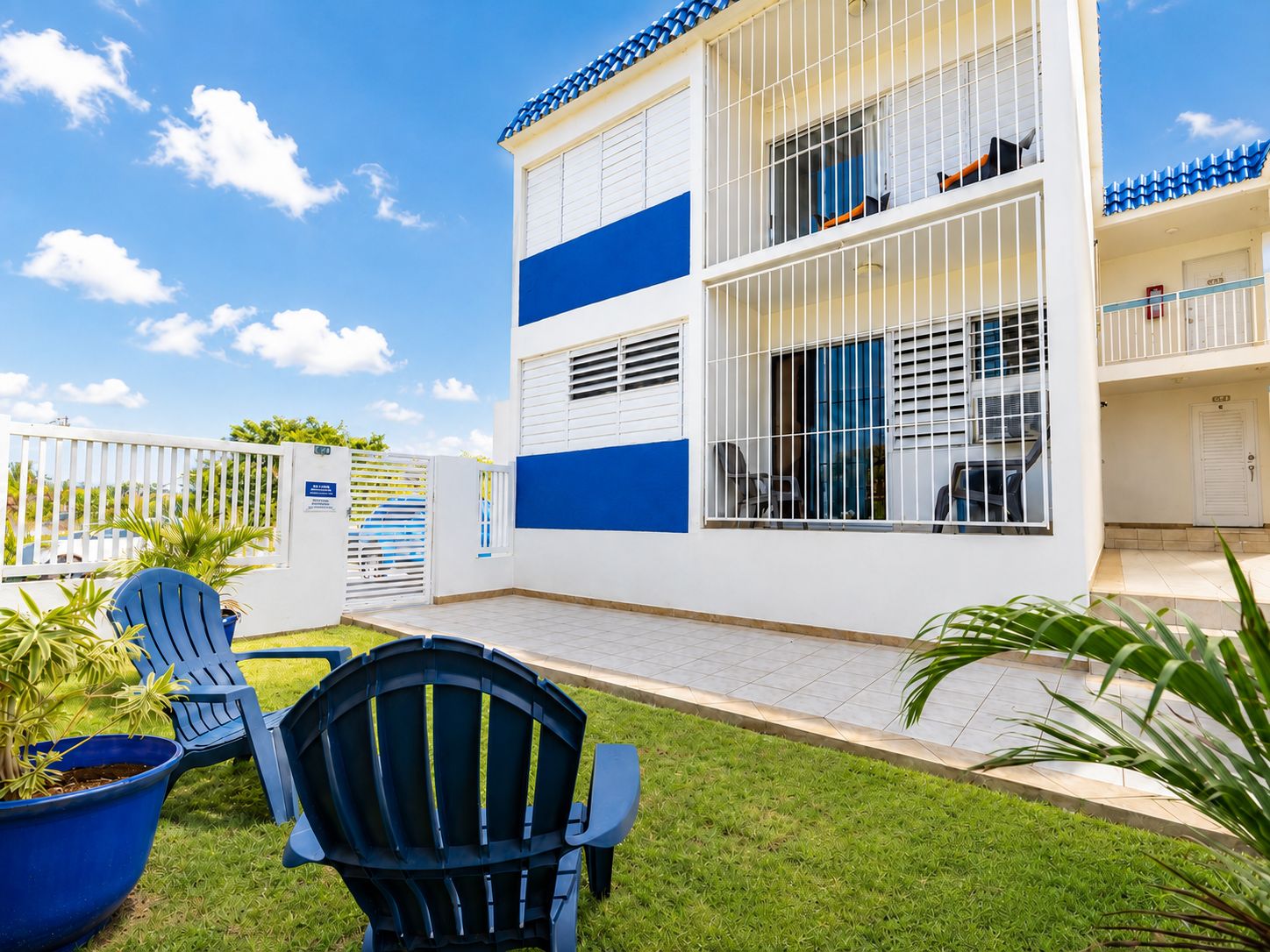 A two-story building with a white and blue facade, a gated entrance, and a small patio area with chairs and potted plants.