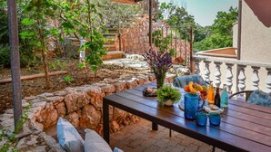 Patio esterno con parete in pietra, tavolo di legno con bevande e snack, e vista su una scala in pietra e vegetazione.