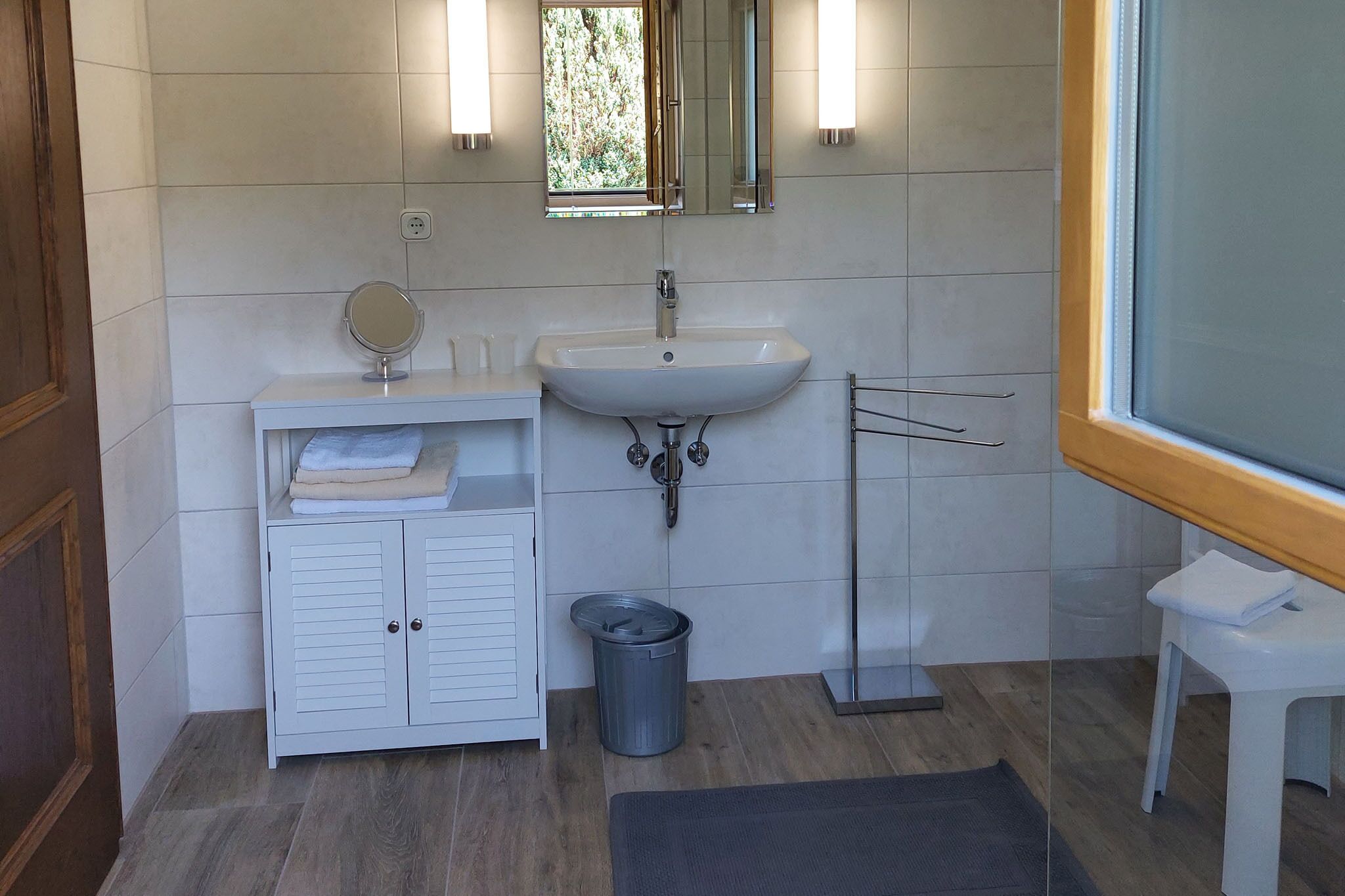 A bathroom with a white vanity, a sink, a mirror, a trash bin, and a window.