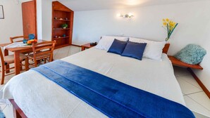 A bedroom with a bed, bedside table, and a small desk with a chair. There is a wooden wardrobe and a shelf with decorative items.