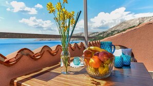 A table with a clear view of the sea, a vase with yellow flowers, and a bowl of fruit.