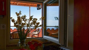 A room with a window overlooking a red-roofed area, a vase with yellow flowers, and a table with a bowl of fruit.