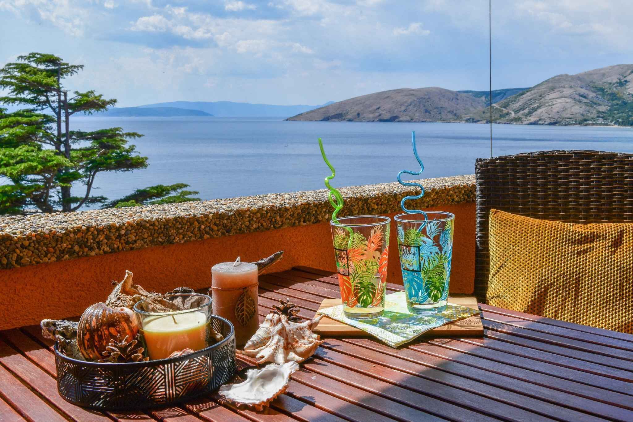 Een houten tafel met drankjes, een kaars en schelpen op een balkon met uitzicht op water en heuvels.