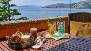 Une table en bois avec des boissons, une bougie et des coquillages, sur un balcon donnant sur une étendue d’eau et des collines.