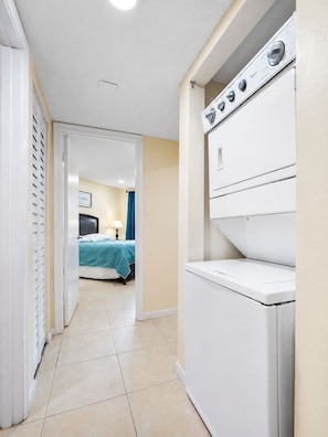 Lavanderia com máquina de lavar e secadora, uma cama no quarto vizinho e uma porta que dá para outra área.