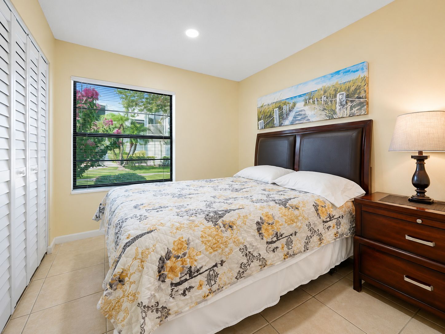 A bedroom with a bed, a nightstand, a lamp, a window with blinds, and a painting on the wall.