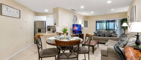 Una sala de estar moderna con sofá, una mesa de comedor con sillas, una zona de cocina con refrigerador y un televisor de pantalla plana grande.