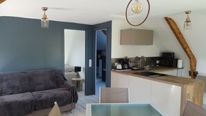 Un salon moderne avec un canapé gris, une petite table ronde avec une lampe et un coin cuisine doté d’un plan de travail en bois et d’une cuisinière intégrée.