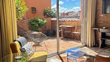 A modern living room with a glass table, a sofa, and a glass coffee table. There is a view of a brick wall and a balcony with outdoor furniture.