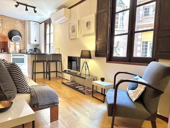 Una sala de estar acogedora con sofá, sillón y mesa de centro, una zona de cocina con radiador y calentador de agua, y amplias ventanas con vistas a edificios.