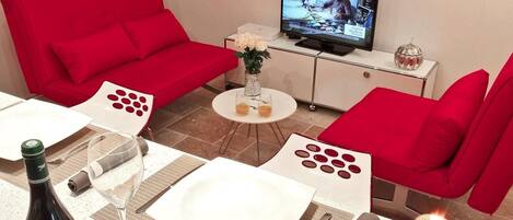 A modern living room with red sofas, a television, a dining table set with wine, roses, and a candle.