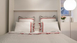 Ein Schlafzimmer mit einem Bett, zwei Kissen, einem Nachttisch, einer Vase mit Blumen und einem Fenster.