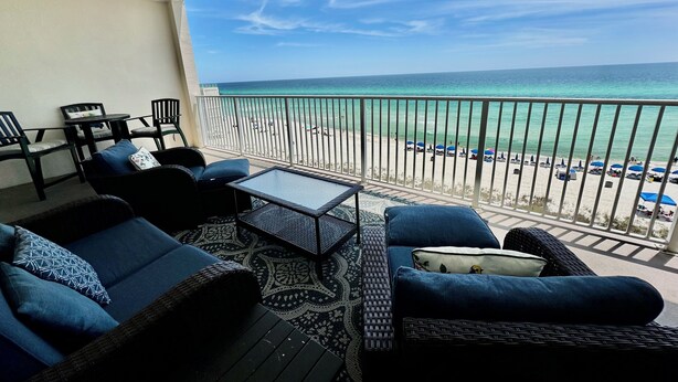 Ein Balkon mit Korbmöbeln, einem Glastisch und Blick auf Strand und Meer.