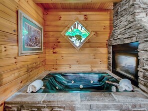 A wooden cabin interior with a stone fireplace, a wooden bathtub with a green marble-like interior, and a stained glass window depicting a green landscape.