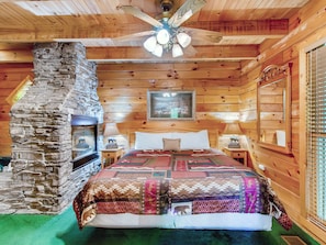 A wooden cabin bedroom with a stone fireplace, a bed with patterned bedding, a ceiling fan, and a framed picture on the wall. - Pet-Friendly | Hot Tub | Secluded | Jacuzzi (Sevierville)