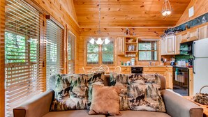 Un interior de cabaña acogedor, con techo de madera, un sofá con diseño de bosque y una zona de cocina con armarios y electrodomésticos de madera.