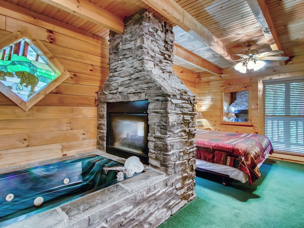 A cozy cabin interior with a stone fireplace, a glass-tub hot tub, and a bed with a patterned blanket.