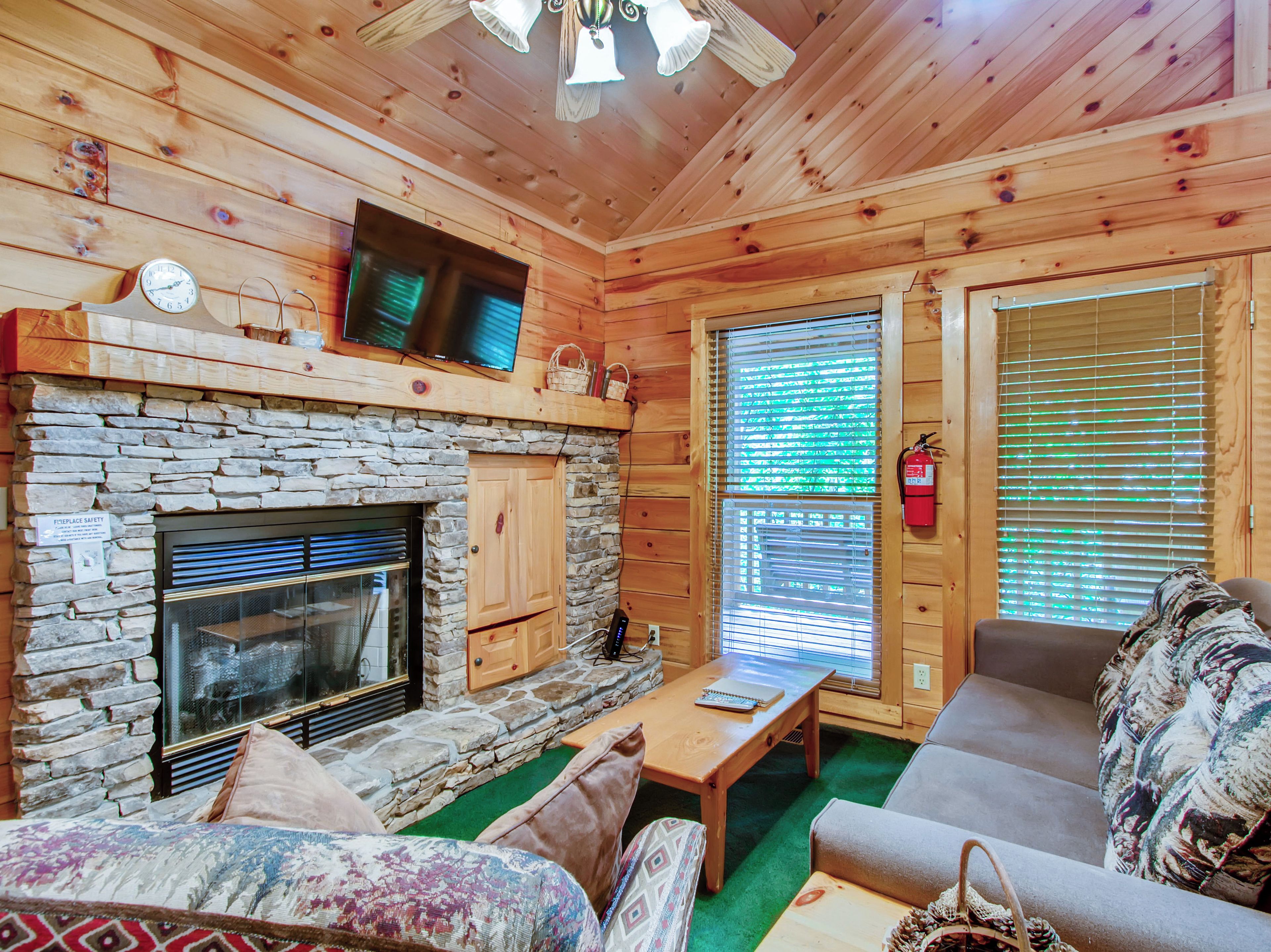 A log cabin interior with a stone fireplace, a television mounted above it, and a sofa with patterned cushions.