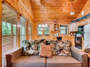 A cozy cabin interior with a wooden ceiling, a sofa with a forest-themed cover, and a kitchen area with wooden cabinets and appliances.