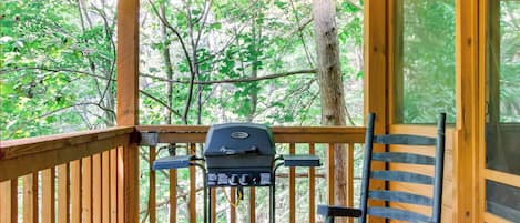 A wooden deck with a grill, a rocking chair, and a view of the forest.