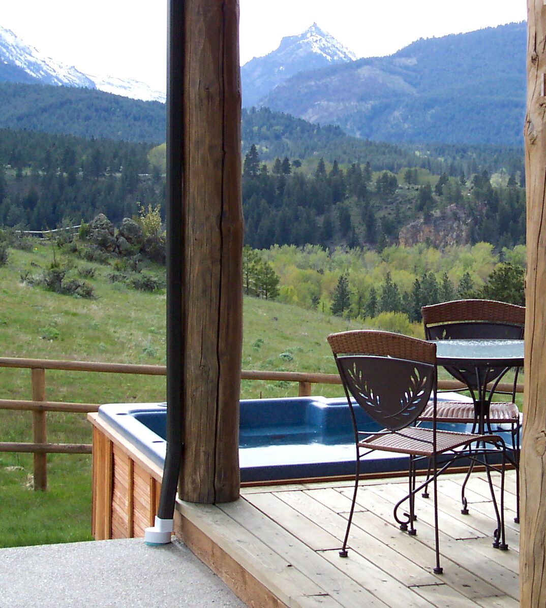 Une terrasse en bois avec un jacuzzi, des fauteuils en osier et une table offrant une vue sur un paysage montagneux.