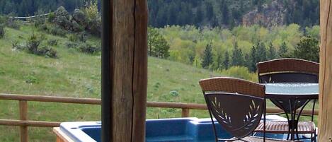 Une terrasse en bois avec un jacuzzi, des fauteuils en osier et une table offrant une vue sur un paysage montagneux.