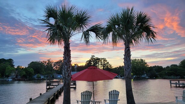 A lakeside view with two palm trees, a red umbrella, and two Adirondack chairs.