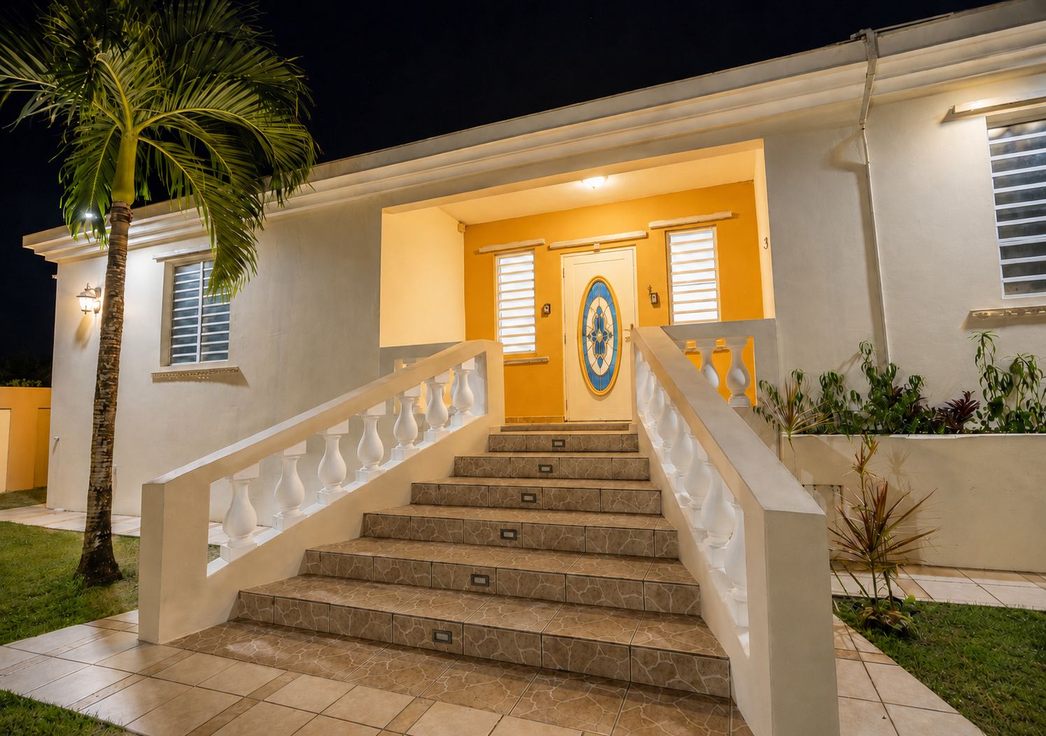 A well-lit entrance with a staircase leading to a door, surrounded by a small garden and a palm tree.