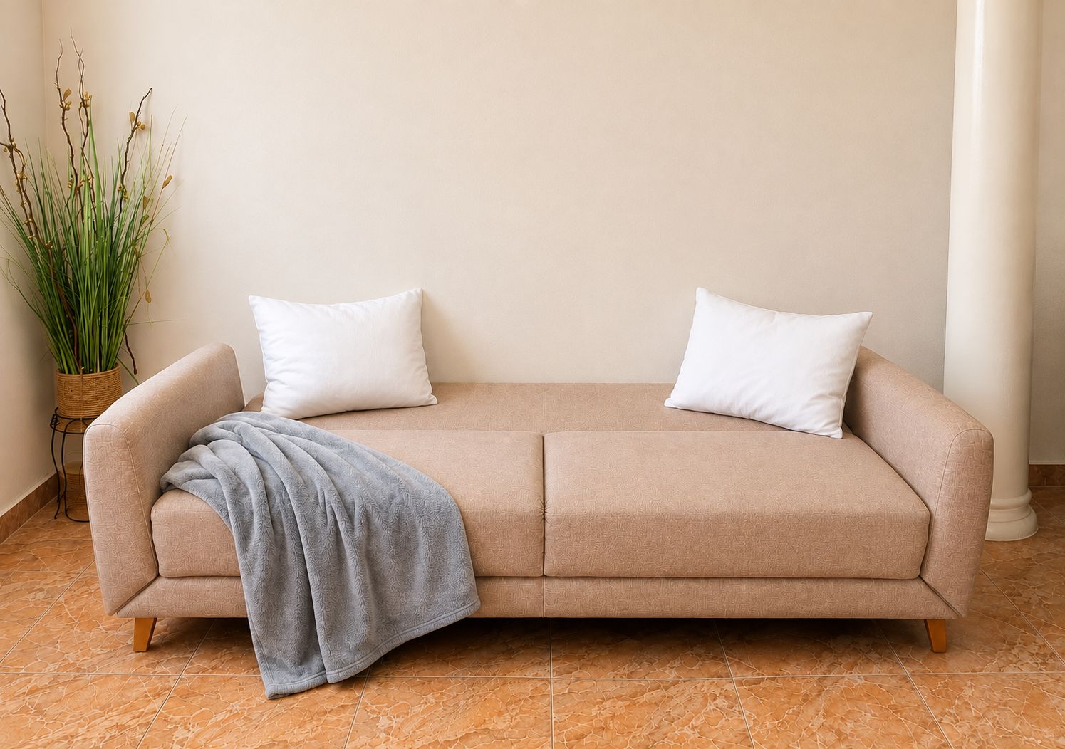 En beige sofa med to hvide puder og et gråt plaide, placeret op ad en ensfarvet væg, med en potteplante til venstre.