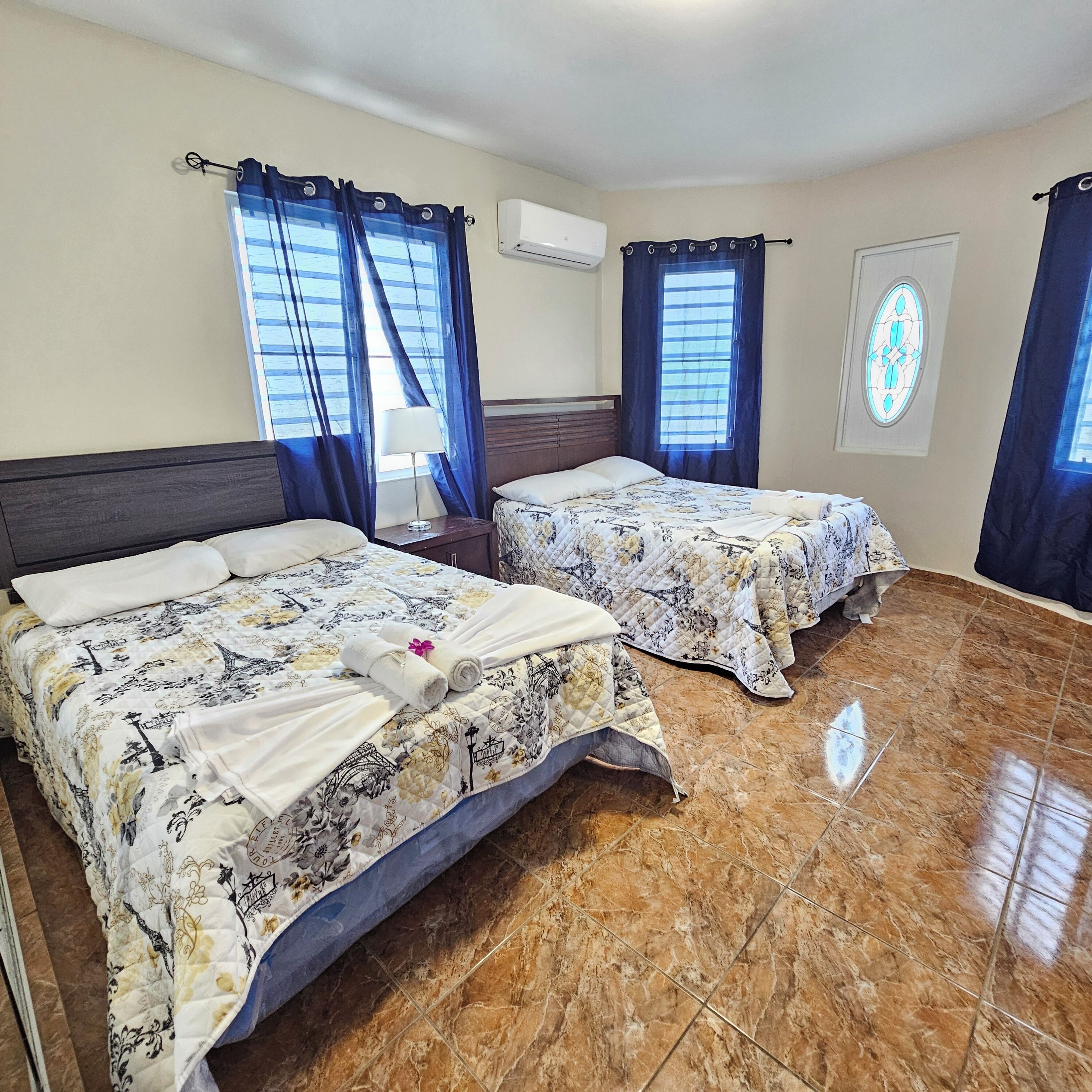 Une chambre d’hôtel avec deux lits, une fenêtre avec des rideaux, un appareil de climatisation et un vitrail.