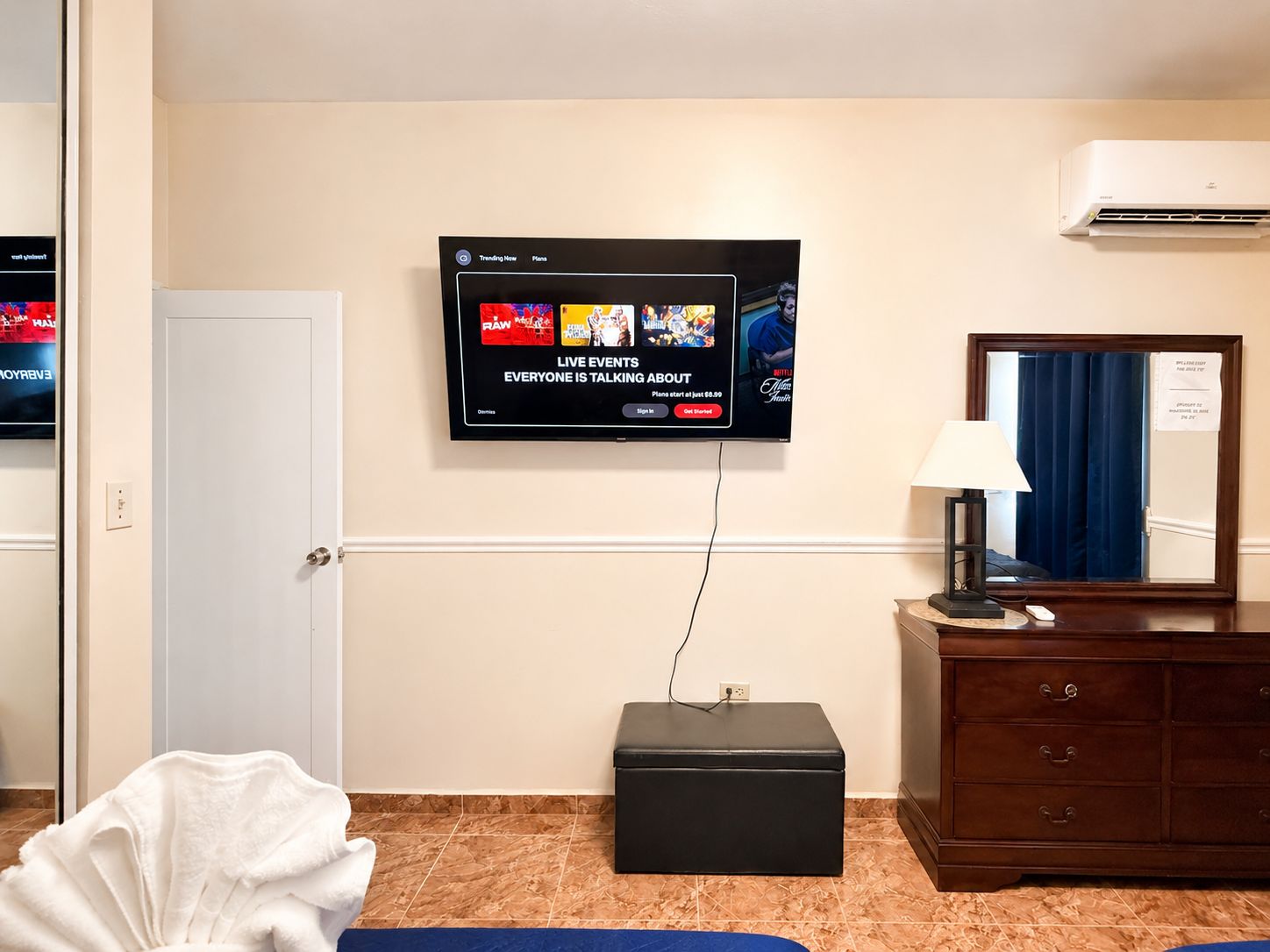 Ein Hotelzimmer mit an der Wand montiertem Fernseher, der eine Live-Übertragung zeigt, einem kleinen schwarzen Hocker, einer hölzernen Kommode mit Schubladen und einem blauen Vorhang.