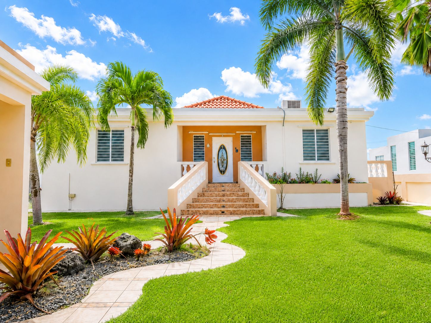 A two-story house with a front entrance, a staircase, and a well-maintained lawn with tropical plants.