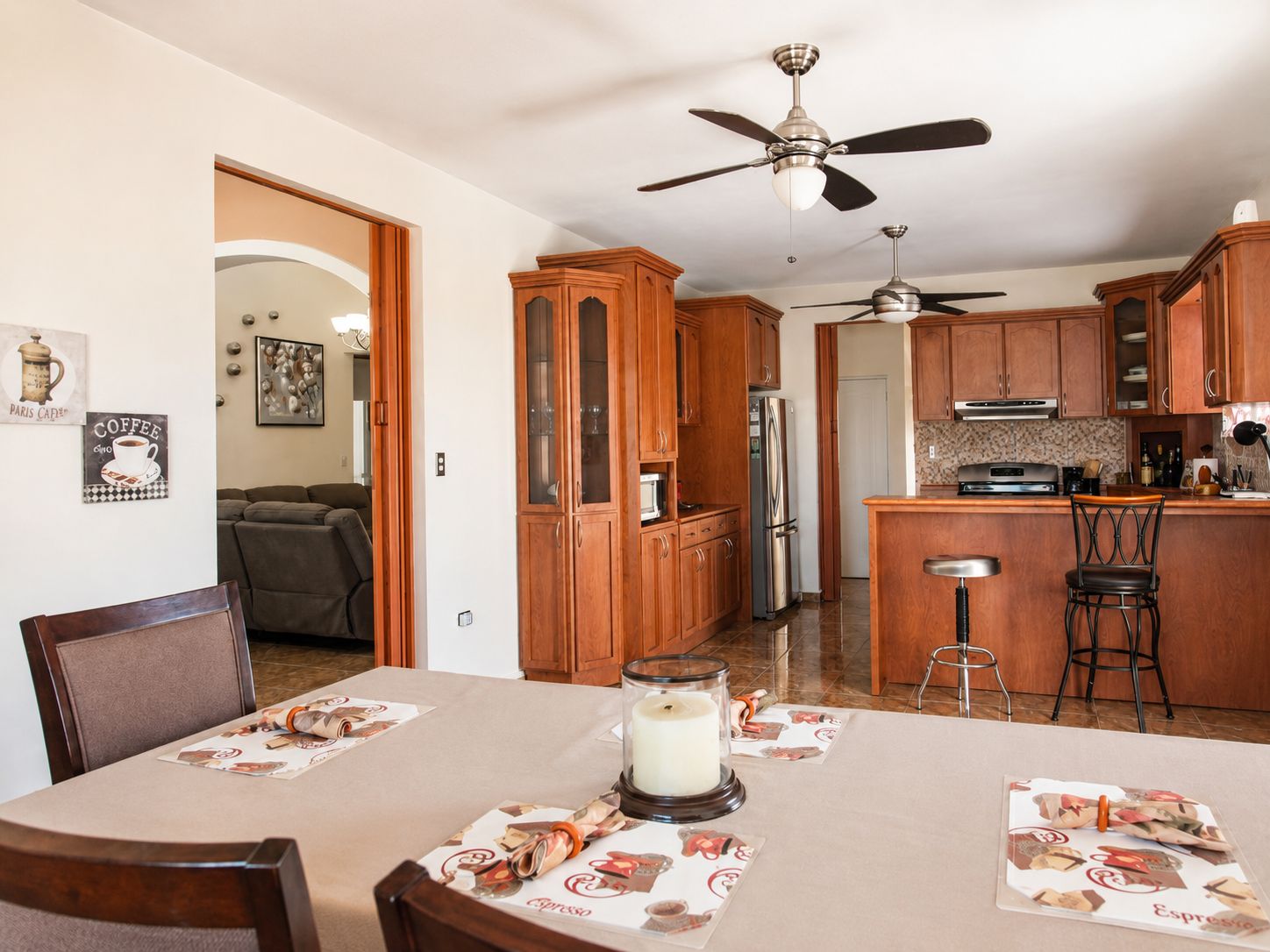Un coin repas avec une table dressée pour quatre personnes, un ventilateur de plafond, des meubles de rangement en bois et une cuisine en arrière-plan.