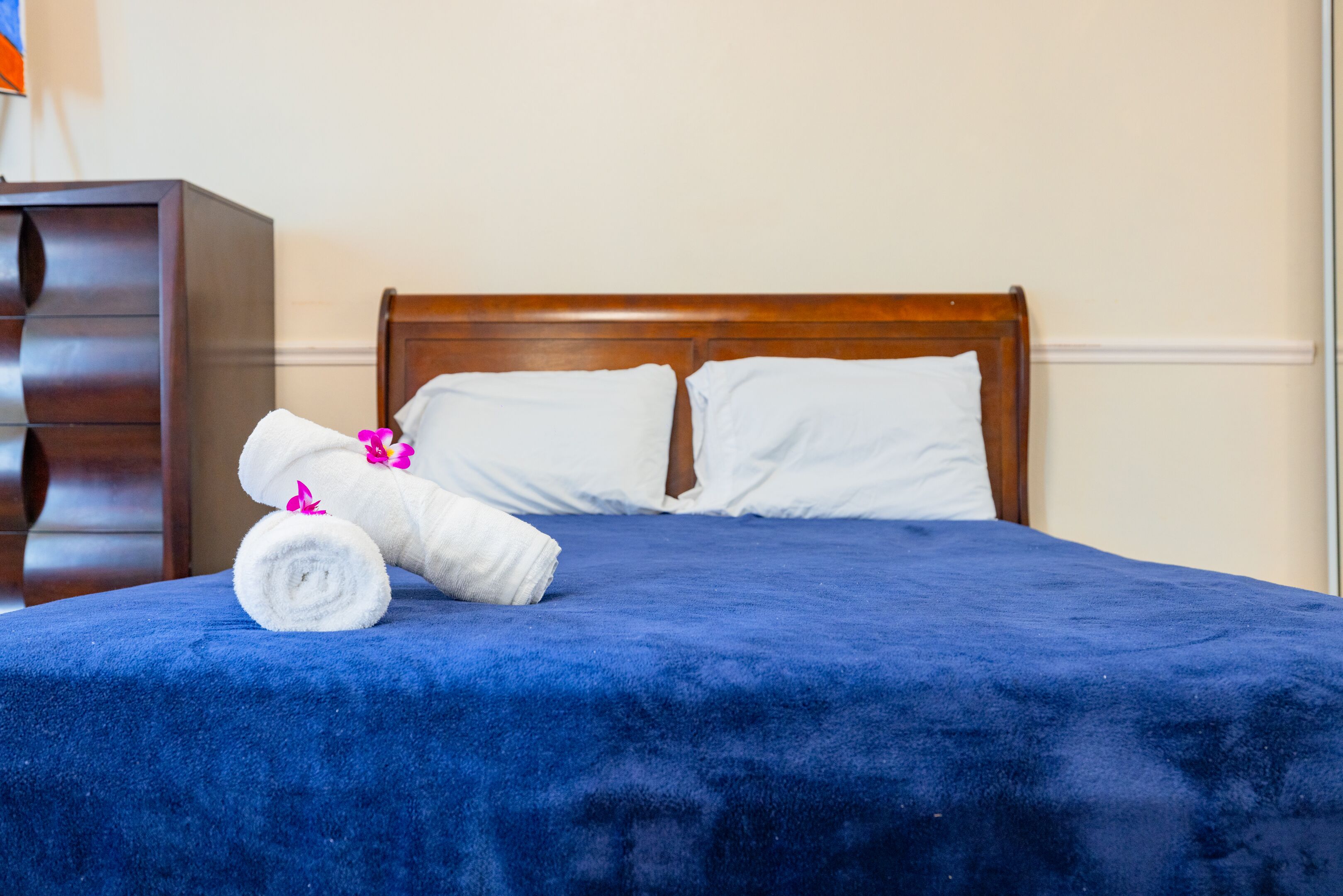 Una cama bien hecha con una manta azul y almohadas blancas, un cabecero de madera y una mesita de noche con una toalla blanca enrollada y flores rosadas.