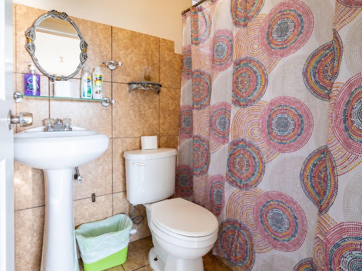 A bathroom with a white sink, a toilet, a green trash bin, a mirror, and a shower curtain with a circular pattern.