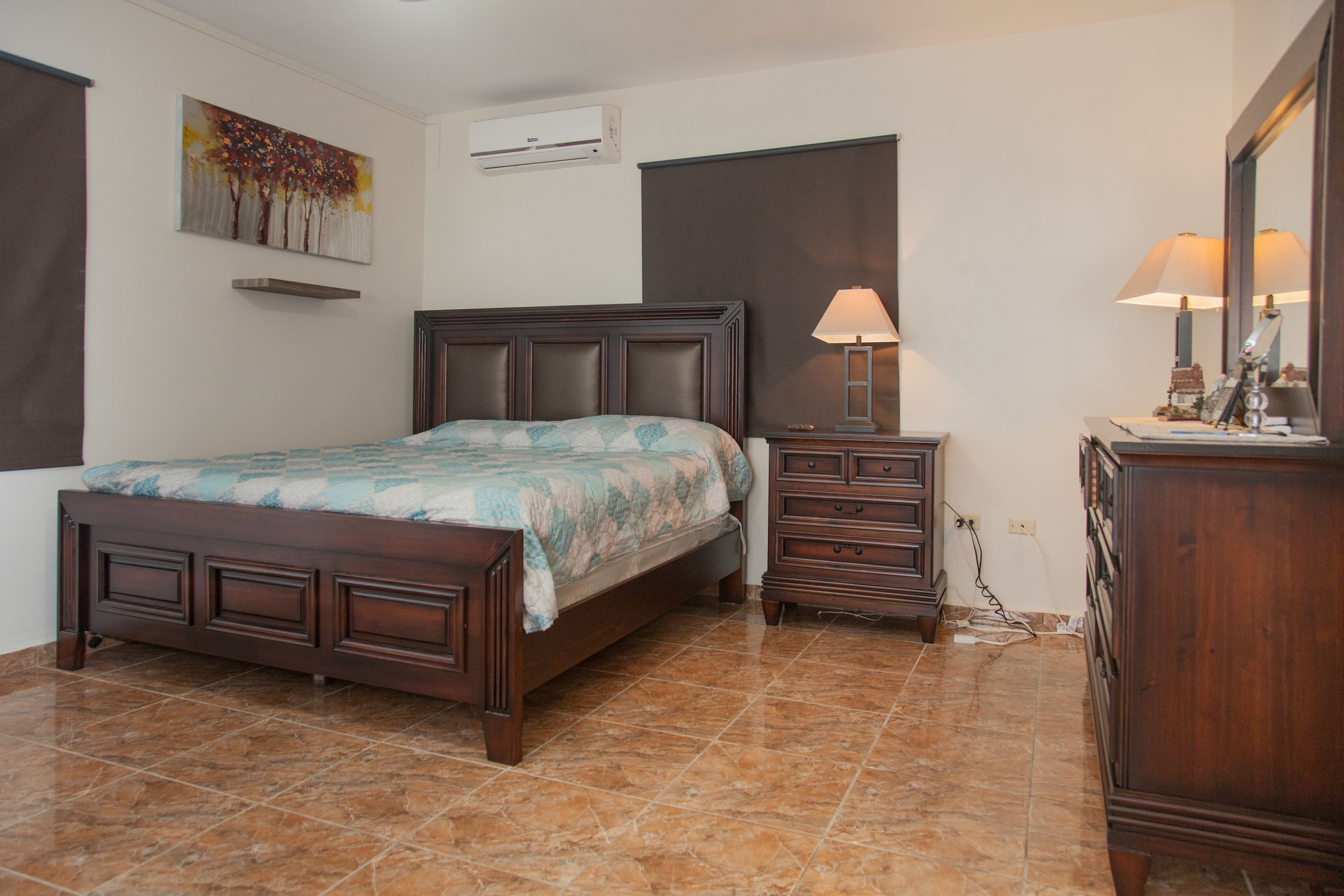 A bedroom with a wooden bed, a nightstand with a lamp, a dresser, and a painting on the wall.
