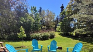 A group of blue Adirondack chairs arranged around a circular fire pit in a grassy outdoor area with trees in the background. - Port Clyde Village House   -   Summer of 2026 is booked. See you in 2027 (St George)