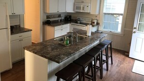 A kitchen with white cabinets, a granite countertop, a microwave, and a sink. There are three stools at the counter. - Port Clyde Village House   -   Summer of 2026 is booked. See you in 2027 (St George)
