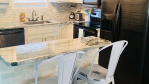 Una cocina con armarios blancos, un refrigerador negro, un microondas y un lavamanos. Hay dos sillas blancas en la isla.