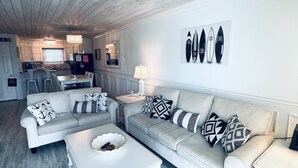 Una sala de estar moderna con un sofá gris, una mesa de centro blanca y cojines estampados. Se ve una cocina al fondo, con una ventana y una araña.