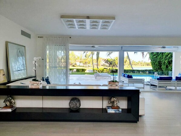A modern living room with a large window, a black console table with decorative items, and a view of a pool and greenery outside.