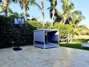 A tent-like structure with a bed inside, a basketball hoop, and palm trees in the background.