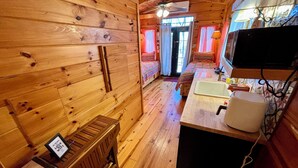 Une cabane chaleureuse avec des murs et des planchers en bois, un lit, une petite kitchenette et un ventilateur de plafond.