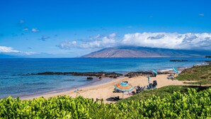 Une plage avec une eau d’un bleu limpide, un rivage rocheux et une montagne en arrière-plan. On y trouve des parasols et des chaises sur le sable.