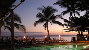 A poolside area with chairs and tables, palm trees, and a view of the ocean at sunset. - ALOHA OceanFront Condo w.A/C,  Molokai, Hawaii Watch whales Dec-April. (Kaunakakai)