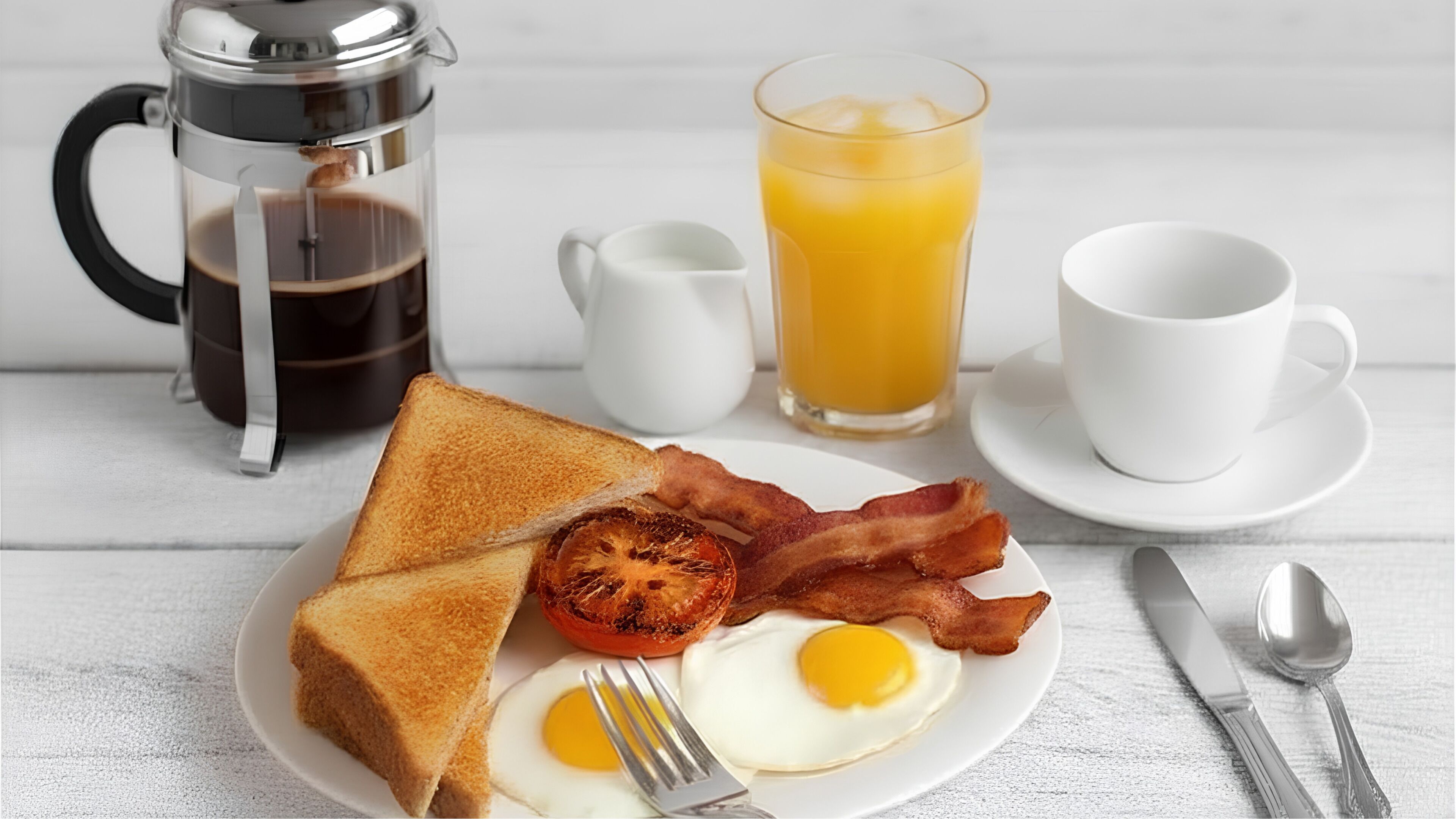 Un plato de desayuno con tostadas, tocino y un huevo, un vaso de jugo de naranja, una cafetera y una taza de café.