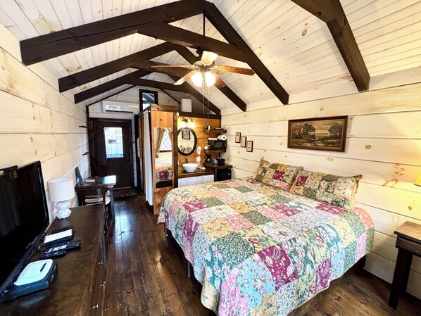 A cozy bedroom with a wooden ceiling, a large bed with a colorful quilt, a bedside table, a nightstand, a desk with a computer, and a window with a view of the outside.