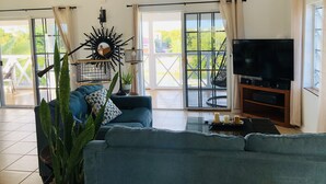A living room with a blue sofa, a ceiling fan, a TV, and a balcony with a view of greenery. - Waterfront Villa, Private Pool & Kayaks (Cooper Jack Bay Settlement)