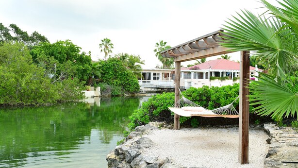 A hammock under a wooden pergola by a calm lake, surrounded by lush greenery and a house with a red roof in the background. - Waterfront Villa, Private Pool & Kayaks (Cooper Jack Bay Settlement)
