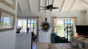 A spacious living area with a high ceiling, large windows, a ceiling fan, a TV, and a sunburst wall decoration. - Waterfront Villa, Private Pool & Kayaks (Cooper Jack Bay Settlement)